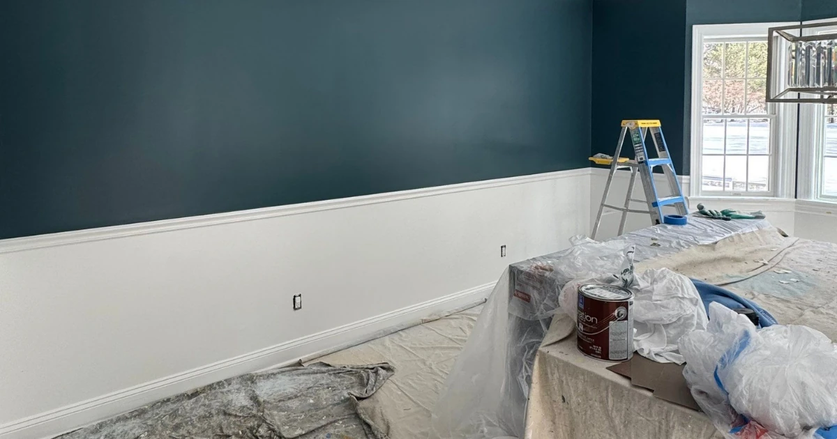 A partially painted room with a dark teal upper wall and white lower wall, a stepladder, and drop cloths covering furniture and a paint can on the floor.