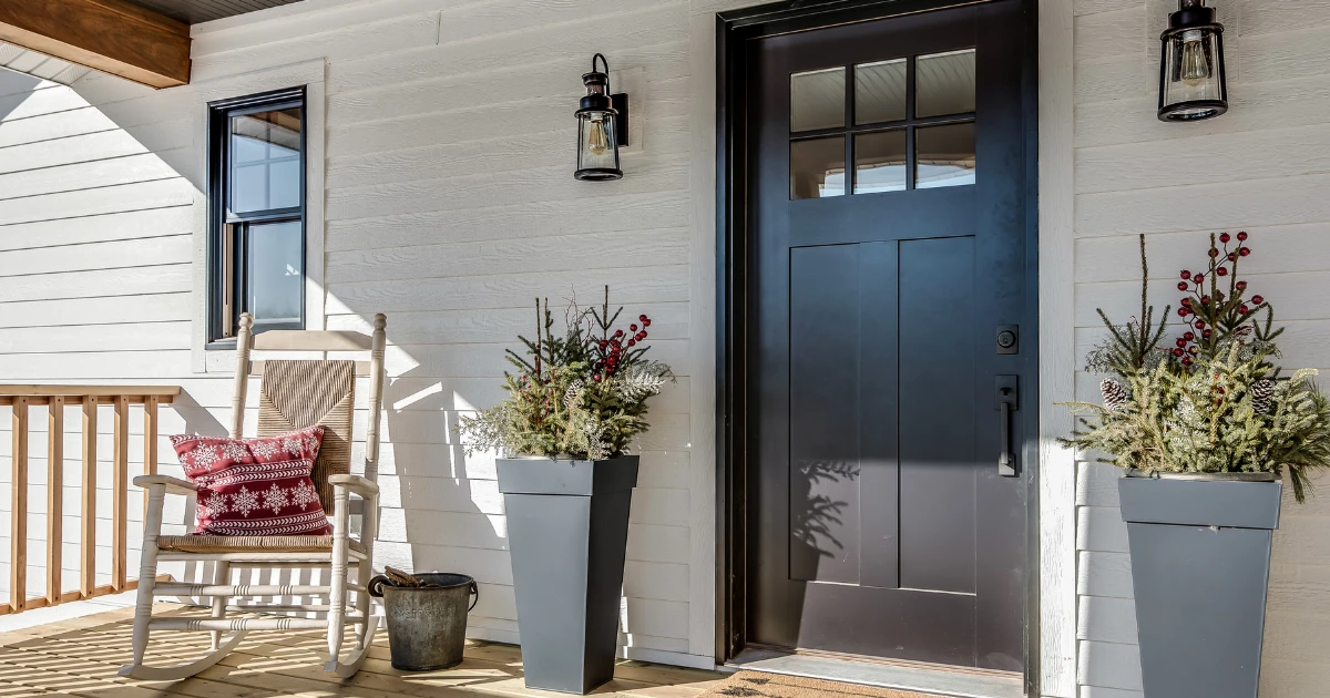 Front porch with white siding, a dark craftsman-style front door, a white rocking chair with a red snowflake pillow, and planters filled with festive greenery.