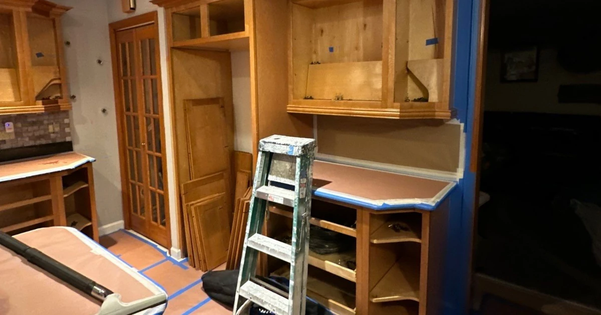 A kitchen prepped for cabinet refinishing, with wood cabinet doors removed and stacked, countertops masked with rosin paper and blue tape, and a stepladder in the center.
