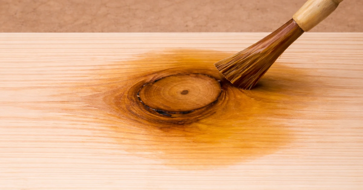 A natural-bristle paintbrush applying amber wood stain over a knot on a light unfinished pine board.