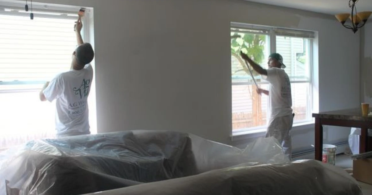 Two painters in white A.G. Williams T-shirts work near windows in a room with furniture covered in plastic sheeting.