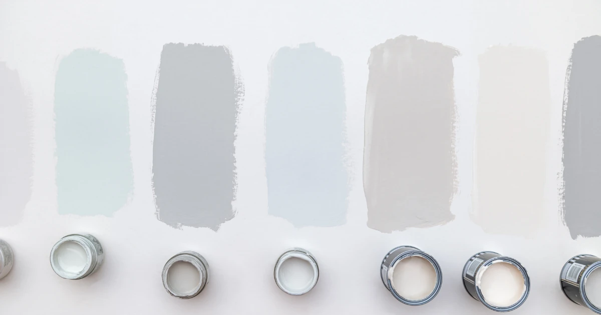 Overhead view of seven paint swatches in soft grays, blues, and beiges brushed onto a white surface, with a row of small open paint cans lined up beneath them.