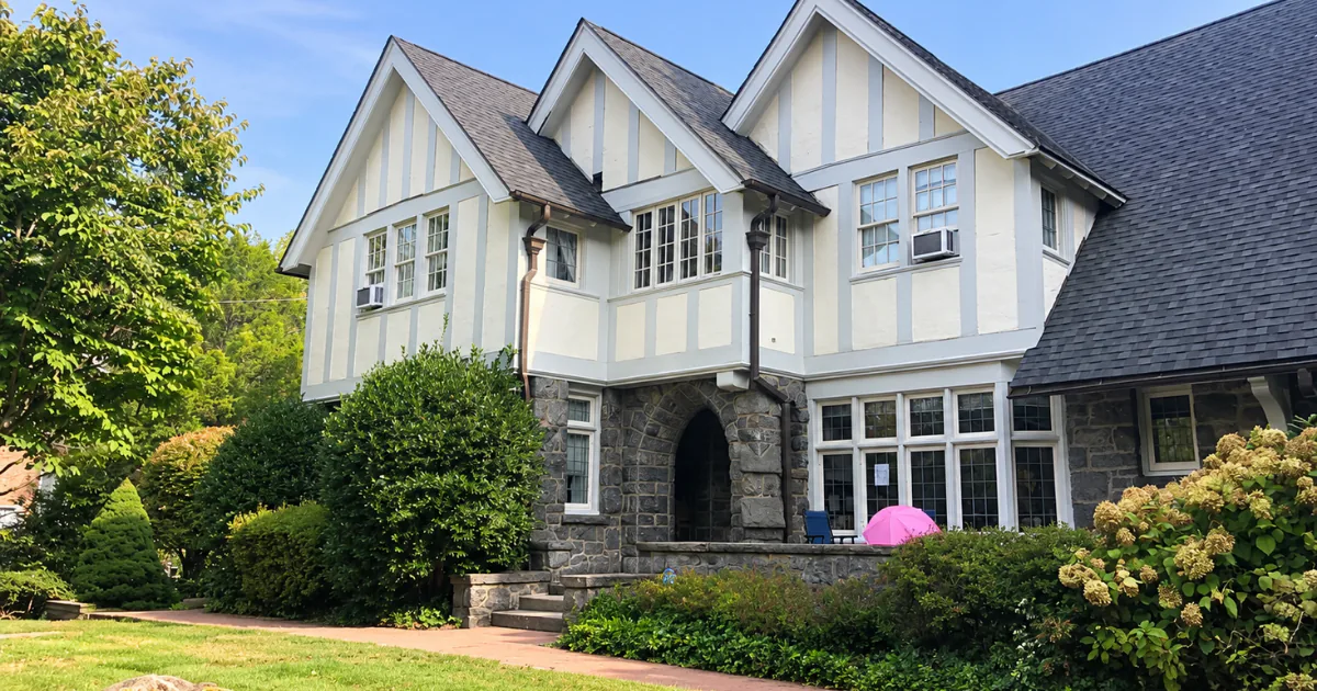 Cream Tudor-style home with gray trim, a stone entry, and a landscaped front yard under a blue sky.
